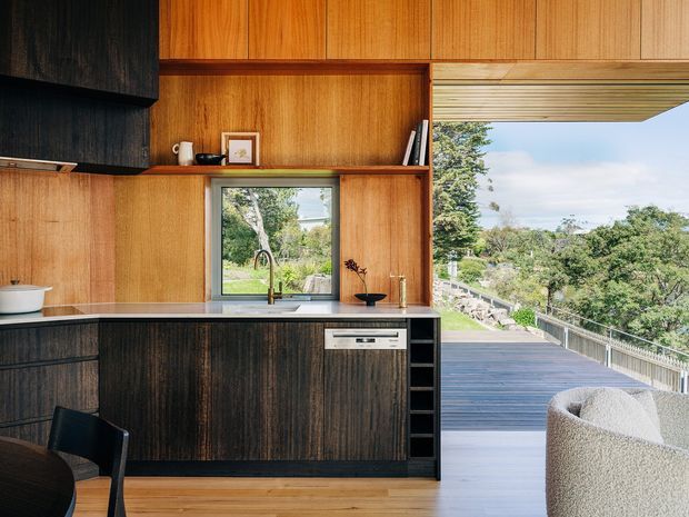 A warm, compact contemporary kitchen with extensive timber wall panelling and cabinetry, dark lower cabinets, a stone benchtop, integrated appliances, and a window above the sink. The kitchen opens directly onto a broad timber deck and landscaped outlook, with a partially visible curved upholstered chair in the foreground. Natural light and timber finishes create a refined indoor-outdoor connection.