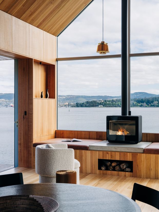 A warm, timber-lined waterfront living and dining space with expansive floor-to-ceiling glazing overlooking a lake and wooded hills. The room features a built-in window seat with upholstered cushions, integrated timber cabinetry and shelving, a contemporary black wood-burning fireplace with log storage, a sculptural pendant light, and a round dining table with upholstered seating. The high sloping timber ceiling and extensive glazing create an airy, refined interior.