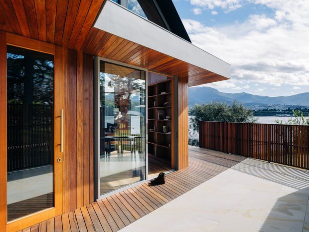 A contemporary timber-clad residential exterior opening onto a spacious elevated terrace. Warm vertical timber wall cladding and soffits surround full-height glazed doors, with integrated dark timber shelving beside the entry. The terrace combines timber decking with a pale tiled or concrete-paved area and is enclosed by a dark vertical-slatted balustrade. Mountainous scenery and water are visible beyond the property.
