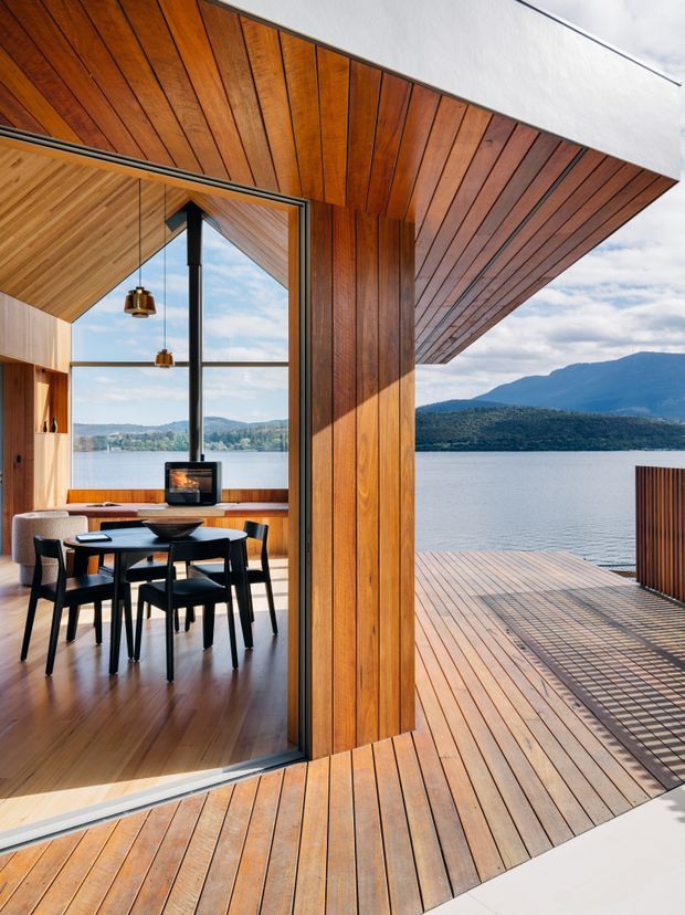 A contemporary lakeside residence viewed from a timber deck, with extensive warm timber wall, ceiling, and floor lining. Large glazed sliding doors open to a dining and living area containing a dark dining table and chairs, a compact fireplace, and pendant lights. The deck projects toward a calm lake with forested mountains beyond, while the dramatic overhanging roof and timber soffit frame the view.