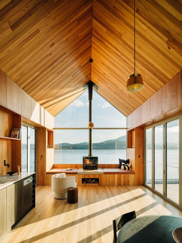 A warm, compact open-plan living space with a dramatic timber-lined vaulted ceiling and matching timber walls and flooring. Floor-to-ceiling glazing frames a calm lake and mountainous landscape. The room includes a built-in window seat, central wood-burning fireplace, compact kitchen cabinetry at left, pendant lighting, and understated contemporary furnishings.