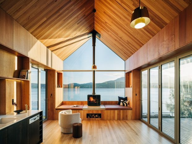 A warm, timber-lined open-plan living space with a compact kitchen at left, extensive glazed sliding doors, and a built-in window seat facing a calm waterfront and mountainous landscape. The dramatic pitched timber ceiling, exposed dark structural post, integrated cabinetry, and pendant lighting create a refined contemporary cabin-like interior.
