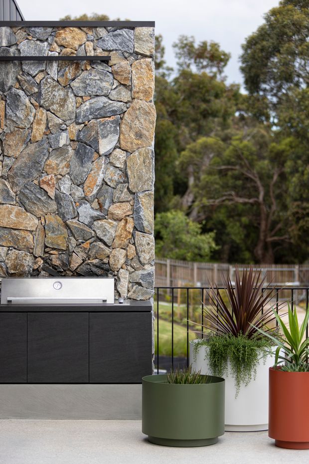 Contemporary outdoor entertaining area featuring a built-in stainless-steel barbecue set into dark cabinetry beneath a dramatic irregular natural-stone wall. The space has a smooth concrete floor, black metal balustrade, and large potted plants, with a leafy landscape visible beyond.