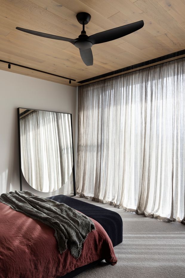 A softly lit contemporary bedroom with a low platform bed dressed in a muted terracotta coverlet and textured charcoal throw. Full-height sheer curtains filter abundant daylight across a large window wall, while a full-height black-framed mirror leans against the light wall. The room features a pale timber-lined ceiling, dark minimalist ceiling fan, black track lighting, and neutral carpet, creating a warm, refined atmosphere.