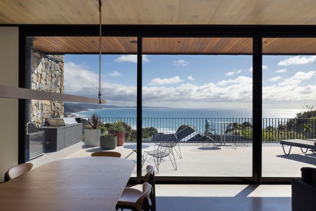 A contemporary open-plan dining and outdoor entertaining space with expansive black-framed glazing overlooking a coastal ocean view. The interior features a light timber ceiling, large timber dining table, and modern pendant light, while the terrace includes an outdoor kitchen, potted plants, dining chairs, sun loungers, and a black metal balustrade. A textured natural stone wall forms a strong architectural feature beside the glazing.