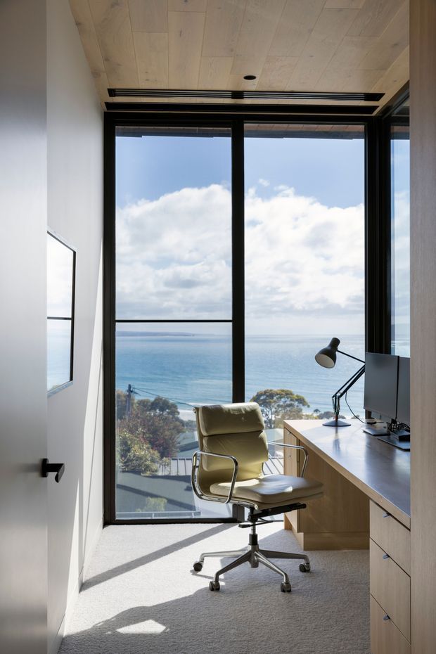 A compact, light-filled home office with a built-in timber desk and drawer storage, ergonomic upholstered office chair, monitor, and angled desk lamp. Full-height black-framed glazing provides expansive ocean views, while the ceiling is clad in pale timber boards and the floor is carpeted in a light neutral tone.
