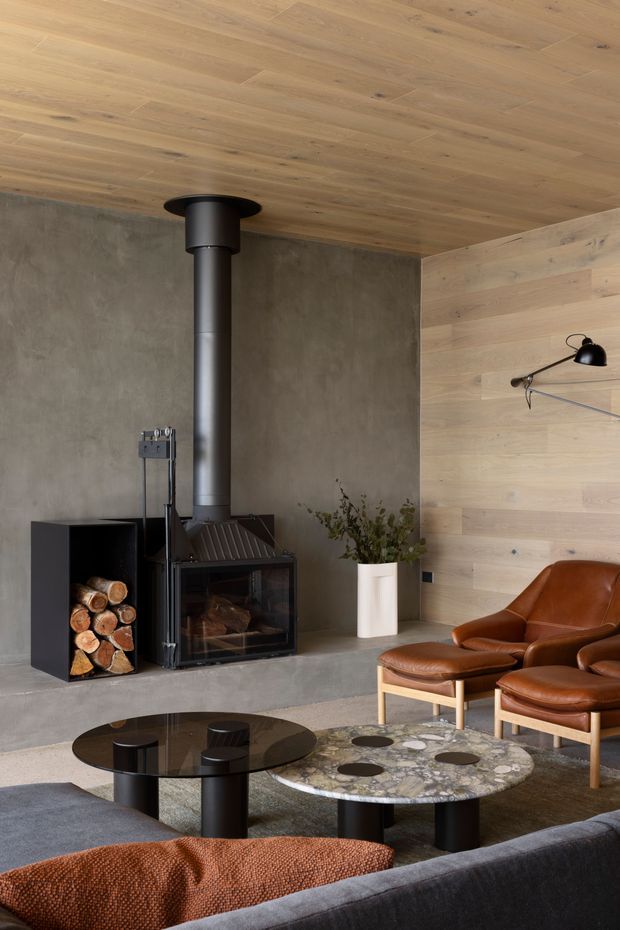 A refined living room centred on a freestanding black wood-burning stove with a tall flue, set against a raw grey concrete wall and light timber-lined ceiling and feature wall. Mid-century leather lounge chairs, a dark glass coffee table, a terrazzo-style table, wool rugs and a low sofa create a warm, sophisticated seating area.