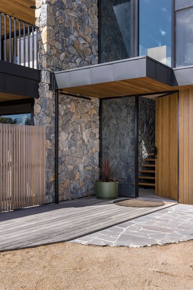 Close-up exterior view of a contemporary residential entry. The facade combines rugged natural stone masonry, dark metal-framed glazing and projecting box forms, with warm timber soffits and vertical timber cladding. A timber deck and irregular stone paving lead to the glazed entrance, with a timber slat privacy screen and a glimpse of an interior stair.