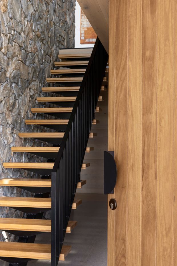 A contemporary stairway with open-riser timber treads, a slim black vertical balustrade, and a dramatic irregular natural-stone wall. Warm timber wall panelling and a concealed door line the right side, with a glimpse of artwork and upper-level space beyond.