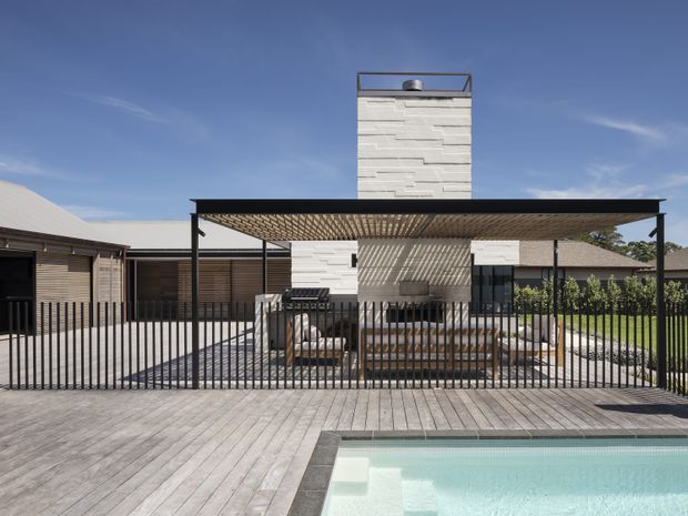 Exterior view of a contemporary single-storey residence with a flat-roofed covered outdoor entertaining area, slender black metal fencing, a pale textured masonry chimney, timber decking, and a swimming pool in the foreground. The restrained linear forms and strong indoor-outdoor connection create a modern architectural composition.