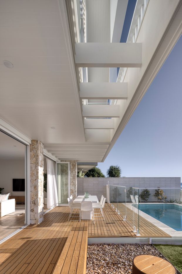 Bright contemporary outdoor terrace beside a swimming pool, viewed from a covered patio. The space features extensive timber decking, frameless glass balustrades, a white outdoor dining setting, stone-clad columns, and a slatted white soffit with overhead clerestory-like glazing. Sliding glass doors open into a light-filled living room, while a rendered boundary wall and clear blue sky form the background.