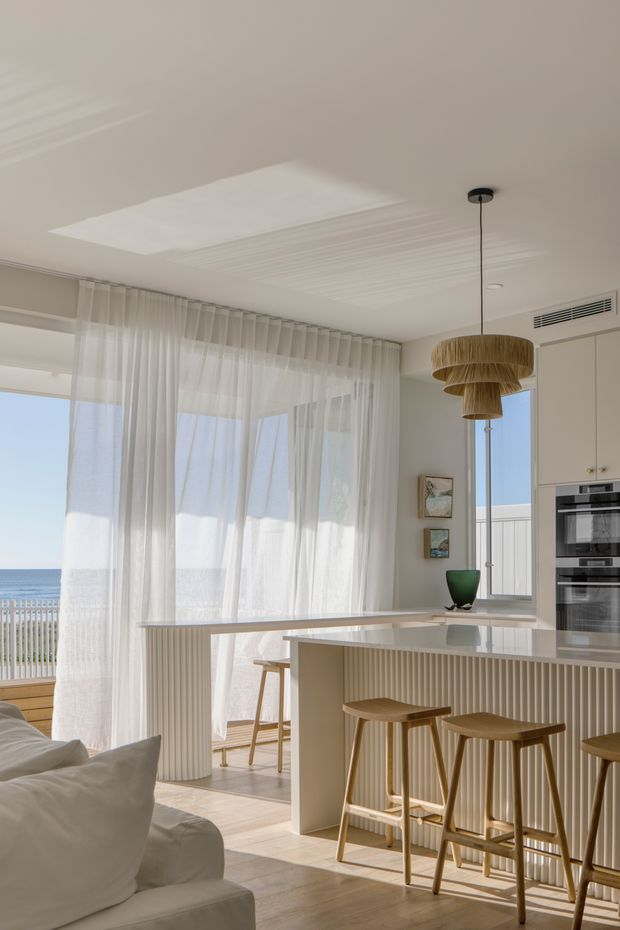 Bright coastal open-plan kitchen and living area with a pale neutral palette, light timber flooring, fluted island cabinetry, integrated appliances, sheer full-height curtains, and a sculptural woven pendant light. The space opens to a balcony with a clear ocean view, creating an airy, relaxed interior.