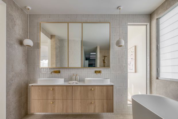 Bright contemporary bathroom featuring a double timber vanity with two countertop basins, brass tapware, large framed mirrored cabinets, textured light wall tiles, a freestanding white bathtub, pendant lighting, and a window with white blinds.