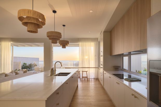 A bright, contemporary open-plan kitchen with a large pale stone island, integrated sink and brass-toned tap, extensive light-wood cabinetry, and timber flooring. Rattan pendant lights hang above the island and dining bench area. Full-height glazing and sheer curtains open to an outdoor terrace with a distant coastal or urban outlook, while a living area is visible to the left.