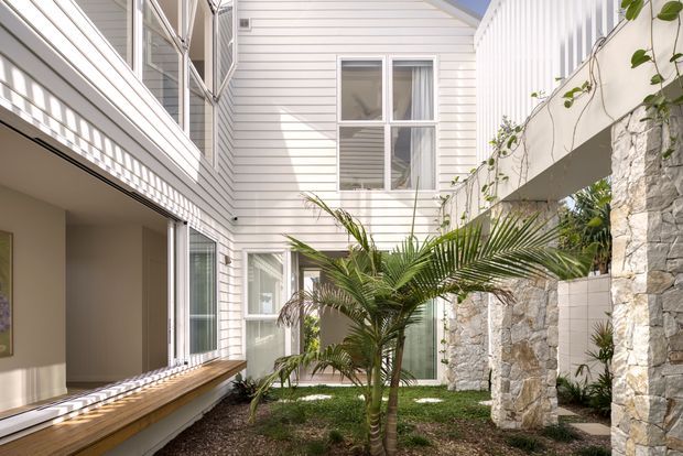 A bright, multi-level internal courtyard framed by white horizontal weatherboard-clad walls, extensive glazing, timber-lined window seating, and pale rough-stone columns. Tropical palms and climbing greenery create a private landscaped garden atmosphere, while strong sunlight and shadow add definition to the contemporary coastal composition.