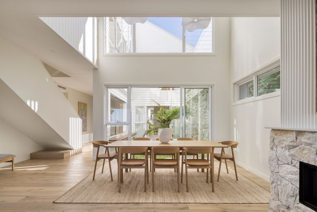 Bright, airy open-plan dining area with a mid-century-inspired timber dining table and chairs set on a large neutral rug. The room features light timber flooring, white walls, extensive glazing, a double-height void, and a stone-clad fireplace with vertical white wall panelling. An adjacent stairway and upper-level windows are visible, creating a spacious, sunlit interior.