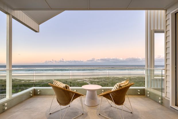 A covered coastal balcony or terrace overlooking the ocean at sunset. The space has a tiled floor, pale timber or painted cladding, slender structural posts, a frameless glass balustrade, and two woven lounge chairs arranged around a small round table. The open-sided design provides expansive beach and sea views.