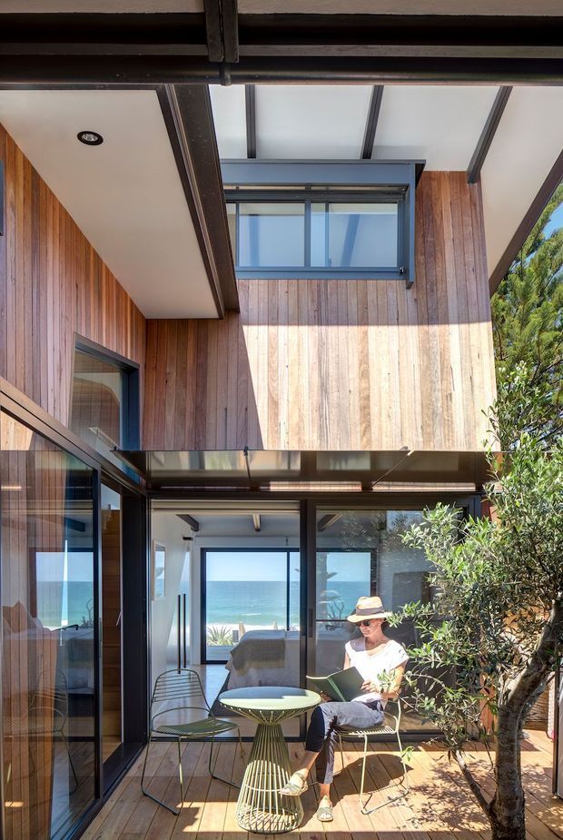 A bright multi-level courtyard-style coastal home exterior with warm vertical timber cladding, dark metal-framed glazing, and a translucent roof canopy. The sheltered outdoor sitting area has timber decking, a small round table, wire-frame chairs, and a potted tree. Through the glazing, an interior bedroom or living space is visible, with a clear ocean view beyond.