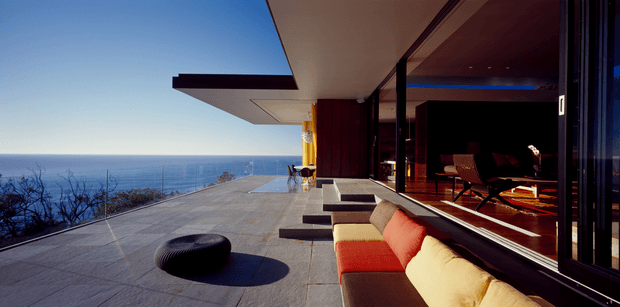 A modern coastal residence with a broad paved terrace overlooking the ocean. The image looks along a cantilevered flat-roofed structure with extensive floor-to-ceiling glazing, dark framed sliding doors, and a partially visible warm-toned interior living area. Outdoor seating and a low round ottoman occupy the terrace, while the architecture emphasizes strong horizontal lines and indoor-outdoor connection.