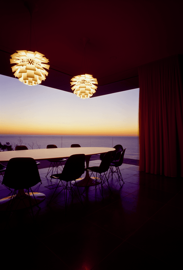 A dimly lit dining room opening onto a broad ocean view at sunset. A long pale dining table is surrounded by dark sculptural wire-base chairs, with two illuminated layered pendant lights hanging from a dark ceiling. Full-height dark curtains frame the glazing on the right, while large dark floor tiles reflect the warm pendant light and sunset colours. The scene has a dramatic, refined mid-century-inspired atmosphere.