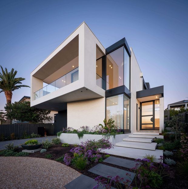 Contemporary two-storey residential facade with bold geometric forms, a projecting upper-level balcony, extensive floor-to-ceiling glazing, and a recessed entry. The exterior combines white rendered walls, dark cladding and framing, warm interior lighting, and landscaped front garden beds with stepping-stone paths.