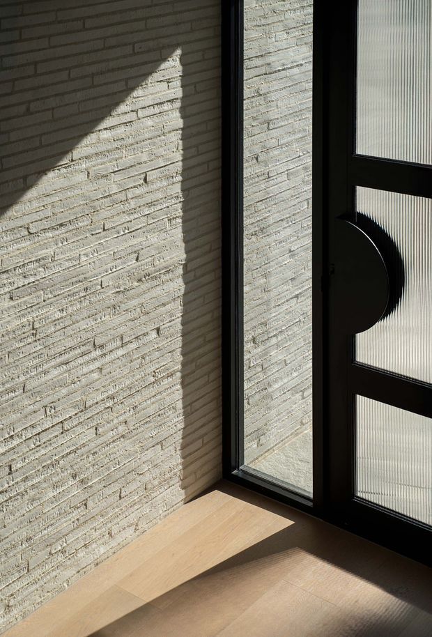 A close-up architectural interior detail showing a pale textured brick wall, warm light timber flooring, and a black-framed glazed door or window with ribbed translucent glass and a distinctive circular dark handle. Strong sunlight creates geometric shadows across the wall and floor.