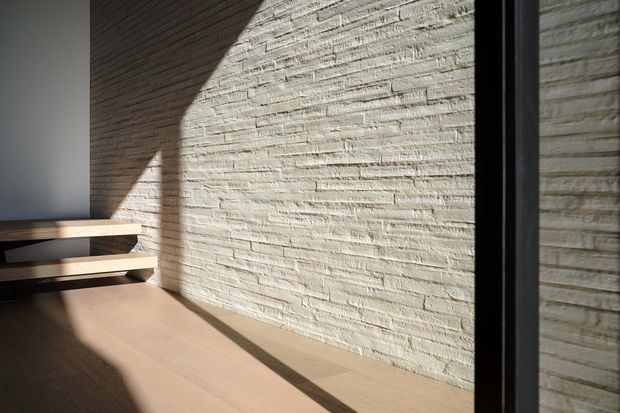 A minimalist architectural interior detail showing a pale textured brick feature wall illuminated by strong sunlight and shadow. A simple floating timber stair or bench-like structure appears at left, with warm timber flooring and a dark-framed glazed opening or partition at right.
