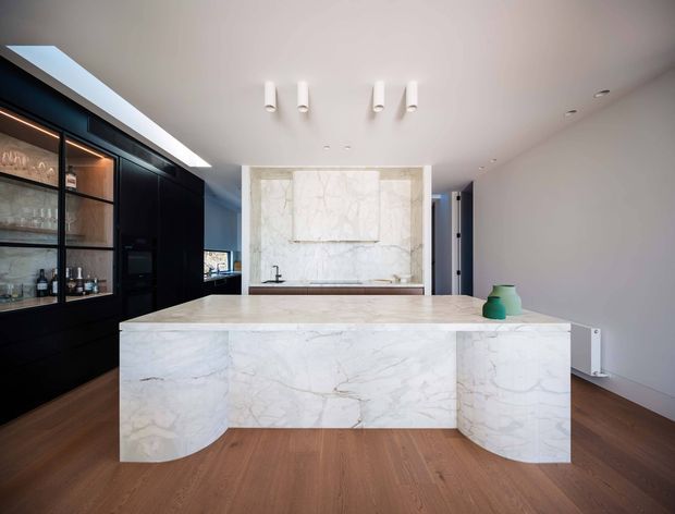 Contemporary open-plan kitchen featuring a large sculptural island clad in white veined stone with rounded ends, dark black cabinetry and glass-fronted display joinery, integrated appliances, a matching stone splashback, warm timber flooring, and a minimalist white ceiling with recessed and surface-mounted lighting. A skylight or clerestory opening brings additional daylight into the space.