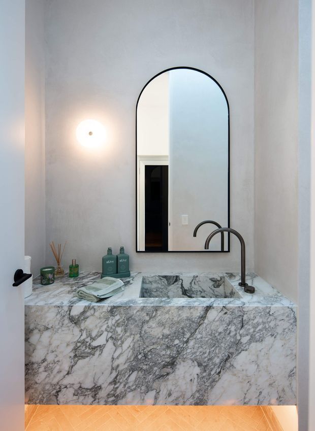 A compact, refined bathroom vanity with a full-height marble-clad basin surround and integrated rectangular sink. The dramatic grey-veined white marble continues across the benchtop and front panel. A tall arched mirror with a slim black frame is mounted above the vanity, alongside a warm globe wall light. Matte black curved tapware and a black wall-mounted toilet paper holder provide contrast. Small toiletries and folded towels sit on the benchtop, while warm timber flooring is visible below.