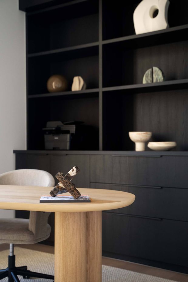 A refined home office or study corner featuring a round light timber table with a pedestal base, a pale upholstered office chair, and dark built-in cabinetry with open display shelving and lower drawers. Neutral sculptural objects, a printer, books, and an abstract sculpture accessorise the shelving and tabletop. The space has a warm, understated, contemporary feel with strong contrast between the dark joinery and light timber furniture.