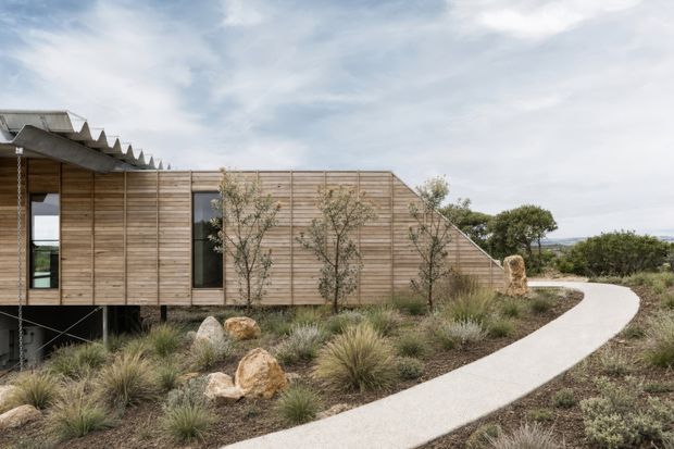 Exterior view of a low, contemporary timber-clad building set within a natural, landscaped site. The structure appears elevated on slim supports, with a sawtooth roof profile and a glazed opening at the left. A pale curved concrete or paved walkway leads through grasses, shrubs and rocks toward the building.