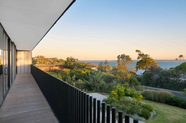A contemporary upper-level balcony or terrace with timber-look decking, dark vertical metal balustrades, and a broad coastal outlook. The image looks across landscaped suburban gardens and rooftops toward blue water, photographed in warm late-afternoon light. The building has clean horizontal lines, a flat soffit, full-height glazing, and dark framed exterior elements.