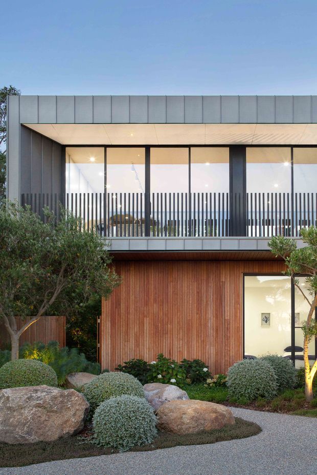 Exterior view of a contemporary two-storey residence with a minimalist rectangular form. The upper level has expansive black-framed glazing, a covered balcony or recessed terrace with vertical metal balustrade elements, and grey metal cladding. The lower level features warm vertical timber cladding and a glazed entrance. A carefully landscaped front garden includes sculpted shrubs, rocks, and a gravel path.
