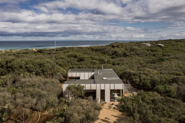 Aerial view of a low-profile contemporary coastal dwelling surrounded by dense native scrub, with the ocean visible beyond. The house has a rectilinear form, flat or very low-pitched grey roofing, light vertical wall cladding, extensive glazing, and a sandy access path leading through the vegetation.