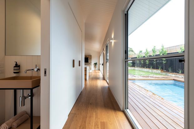 A bright, narrow residential hallway with warm timber flooring, white built-in storage, and full-height glazing opening to a timber deck and swimming pool. A compact wall-mounted vanity with a brown benchtop is visible in the foreground on the left, while the corridor leads toward a light-filled living area.