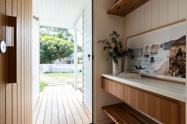 A bright residential entryway viewed from inside, with a timber front door at left and a glazed opening onto a covered timber porch and landscaped yard. The entry features a built-in timber console with open shelving, a pale benchtop, framed coastal artwork, decorative books and a vase of foliage. White vertical wall cladding and a white-lined ceiling create an airy, relaxed coastal character.