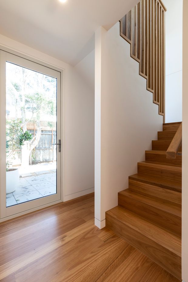 A bright residential stairway and entry area with warm timber flooring and a matching timber staircase. White walls and a full-height glazed exterior door bring in natural light, while a vertical timber-slat balustrade follows the stair profile. The space has a clean, contemporary architectural appearance with simple white detailing and integrated timber handrails.