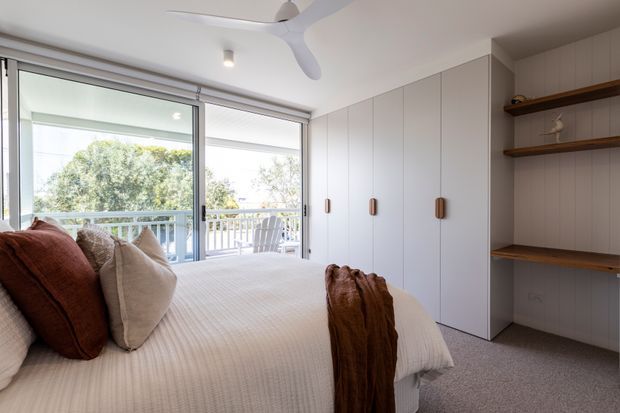 Bright, compact bedroom with a neatly made bed, warm rust and neutral cushions, full-height white built-in wardrobes, and open timber display shelves. Sliding glass doors open to a balcony with leafy views, while a white ceiling fan and soft grey carpet complete the calm contemporary interior.