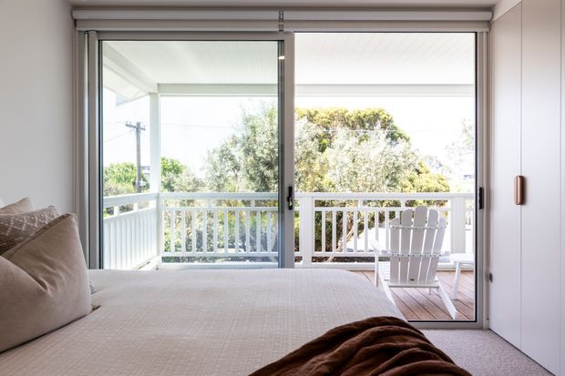 Bright bedroom viewed toward a covered outdoor balcony through wide sliding glass doors. The room has a large upholstered bed with neutral bedding, pale walls, carpeted flooring, built-in white wardrobe cabinetry, and a timber-decked balcony with white railings and outdoor seating beyond.