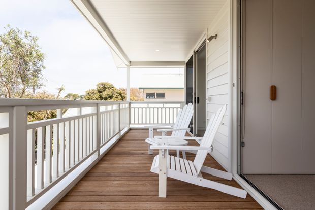 A bright covered timber deck or balcony viewed from beside an open doorway. The space features white-painted horizontal weatherboard walls, a white slatted ceiling, white balustrades, timber decking, and two white Adirondack-style lounge chairs. The elevated outdoor area overlooks trees and a neighbouring building, creating an airy residential setting.