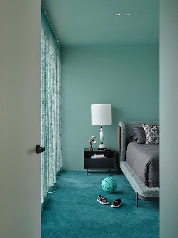 A compact, monochromatic teal bedroom viewed through an open doorway. The room features teal painted walls and ceiling, full-height textured curtains, teal carpet, and a textured upholstered bed with dark grey bedding. A black bedside table with a white lamp sits beside the bed, while a teal basketball and sneakers rest on the floor. Recessed ceiling lights and a dark door handle add simple contemporary details.