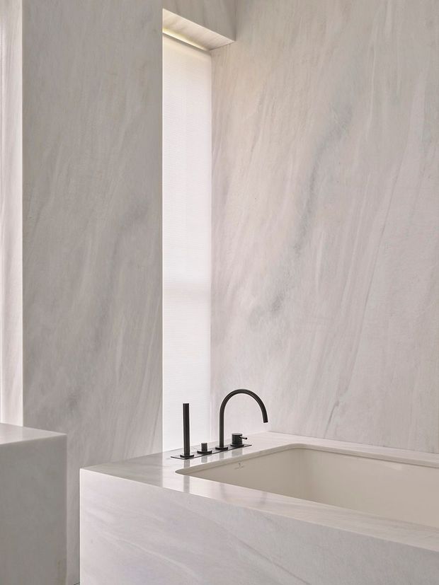 Minimalist bathroom with a freestanding or built-in bathtub clad in pale veined stone, matching stone walls and a crisp white integrated ledge. A matte black arched faucet and separate controls provide contrast, while a narrow window and concealed linear lighting add soft illumination.