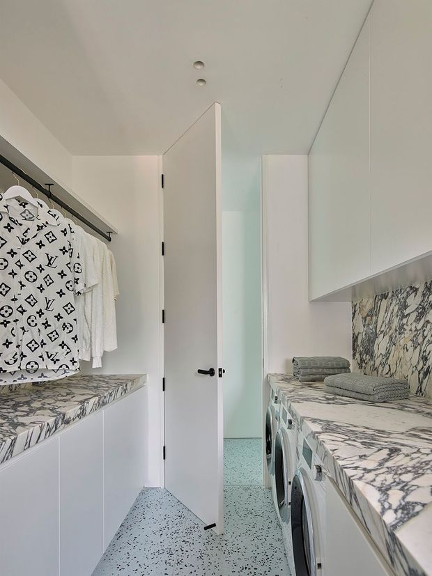 Bright, narrow contemporary laundry room with white cabinetry, a long marble-patterned countertop and matching backsplash, front-loading washing appliances, a terrazzo-style speckled floor, and an open door. Clothes hang from a black rail above the left counter, while folded towels sit on the right.