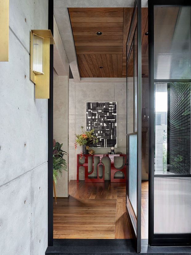 A refined contemporary interior entry or hallway viewed through a dark-framed glazed partition. The space features warm timber flooring and a timber-lined ceiling, large pale concrete or stone-look wall panels, black metal-framed glazing, and a distinctive red console table with sculptural stools beneath monochrome artwork. A brass wall light and indoor plants add warmth and contrast.