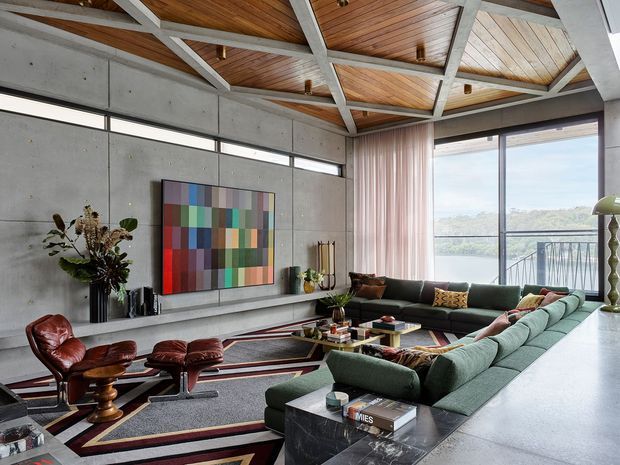 A spacious contemporary living room with a large green sectional sofa, leather lounge chairs and ottomans, a marble-topped coffee table, and a graphic geometric rug. The room features exposed concrete walls, extensive floor-to-ceiling glazing, a timber-lined ceiling with exposed structural beams, and a colourful abstract artwork. A balcony and leafy landscape are visible beyond the windows.