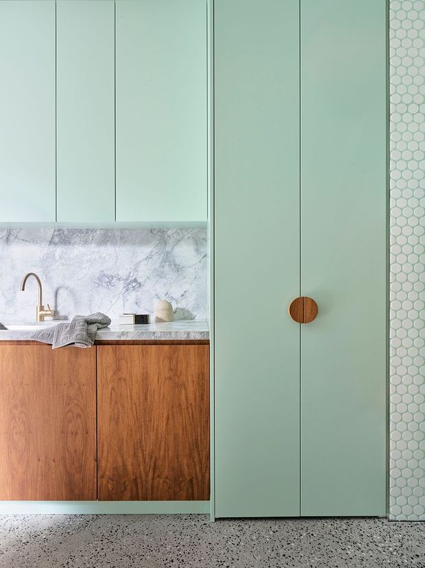A refined contemporary kitchen or kitchenette featuring pale sage-green full-height cabinetry with a round timber pull, warm timber base cabinets, a pale veined stone benchtop and splashback, brushed brass tapware, and a vertical wall of small pale green hexagonal tiles. The floor is a speckled terrazzo-like finish. The composition is tightly framed around the cabinetry and work surface.