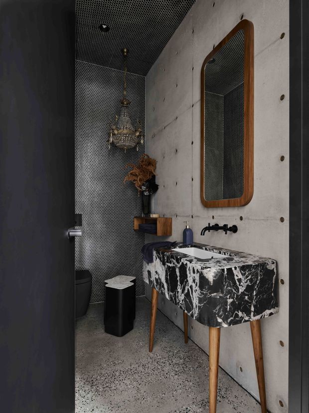 Compact, moody bathroom with a black-and-white marble vanity on timber legs, wall-mounted black tapware, a rounded rectangular timber-framed mirror, exposed concrete wall panels, dark patterned wall and ceiling surfaces, terrazzo flooring, and a decorative pendant light.