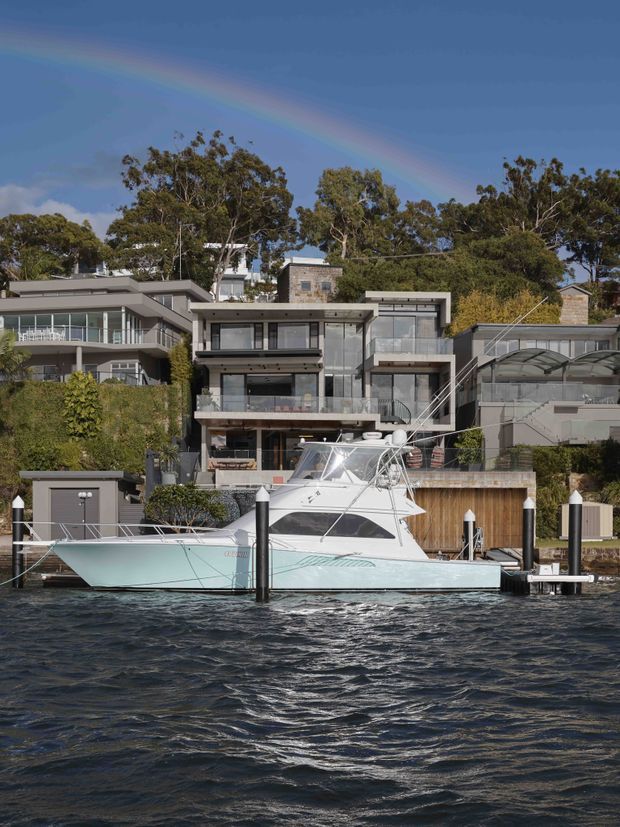 Exterior view of a contemporary multi-level waterfront residence with extensive glazing, balconies, terraces, and a private boat dock. The house is set among mature trees on a landscaped sloping site, with a large yacht in the foreground and a rainbow across the sky.