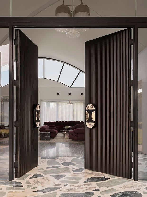View through tall, dark fluted double doors into a refined living room. The room beyond has a curved glazed clerestory or roof window, sheer curtains, sculptural chandelier, deep burgundy sectional seating, and a polished irregular stone floor extending through the doorway. The dramatic entry doors feature narrow vertical ribs and small arched inset windows.