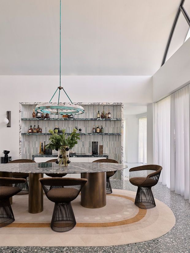 A sophisticated open-plan dining and entertaining space with a large round marble dining table, sculptural wire-frame chairs, and a central floral arrangement. Behind the table is a built-in open bar and display shelving clad in vertically patterned stone or tile, stocked with bottles and glassware. The room features a dramatic high angled ceiling, full-height sheer curtains, terrazzo flooring, a sculptural pendant light, and a large layered circular rug in neutral tones.